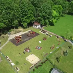 Plac zabaw w otoczeniu zieleni, z huśtawkami, zjeżdżalniami i piaskownicą. W tle budynek i las. To idealne miejsce na wypoczynek dla rodzin z dziećmi w okolicy księża góra radzionków.